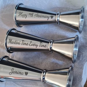 Op de afbeelding: Drie zilveren metalen maatbekers met gepersonaliseerde gravures. De eerste maatbeker zegt "Happy 11th Anniversary" met een hartje. De tweede maatbeker zegt "Martini Time Every Time". De derde maatbeker zegt "Jemima" met een hartje.