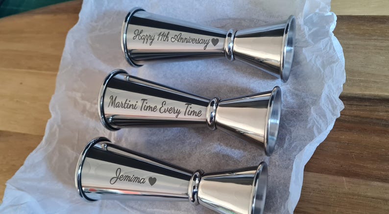 Op de afbeelding: Drie zilveren metalen maatbekers met gepersonaliseerde gravures. De eerste maatbeker zegt "Happy 11th Anniversary" met een hartje. De tweede maatbeker zegt "Martini Time Every Time". De derde maatbeker zegt "Jemima" met een hartje.
