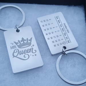 May include: Two silver keychains with engraved designs. One keychain features a crown and the words "His Queen". The other keychain has a calendar with the date "October 2021".