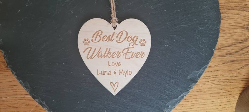 Puede incluir: Un letrero de madera en forma de coraz&oacute;n con el texto "Best Dog Walker Ever" y "Love Luna & Mylo". El letrero tiene dos huellas de patas a cada lado del texto.
