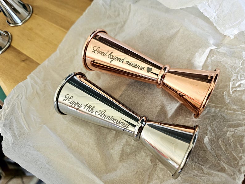 Op de afbeelding: Twee gegraveerde metalen jiggers, een zilveren en een ros&eacute;gouden. De zilveren jigger is gegraveerd met "Happy 11th Anniversary" en de ros&eacute;gouden jigger is gegraveerd met "Loved beyond measure" en een hart.