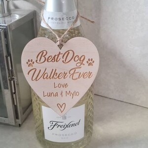 Puede incluir: Un letrero de madera en forma de coraz&oacute;n con el texto "Best Dog Walker Ever" y los nombres "Luna & Mylo" en una fuente m&aacute;s peque&ntilde;a. El letrero est&aacute; colgado de una botella de Freixenet Prosecco.