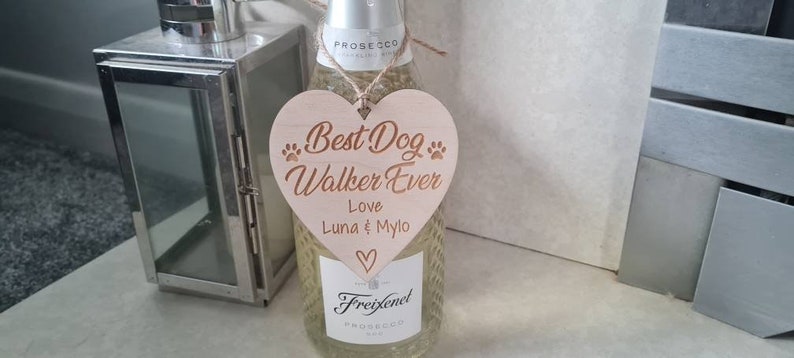 Puede incluir: Un letrero de madera en forma de coraz&oacute;n con el texto "Best Dog Walker Ever" y los nombres "Luna & Mylo" en una fuente m&aacute;s peque&ntilde;a. El letrero est&aacute; colgado de una botella de Freixenet Prosecco.