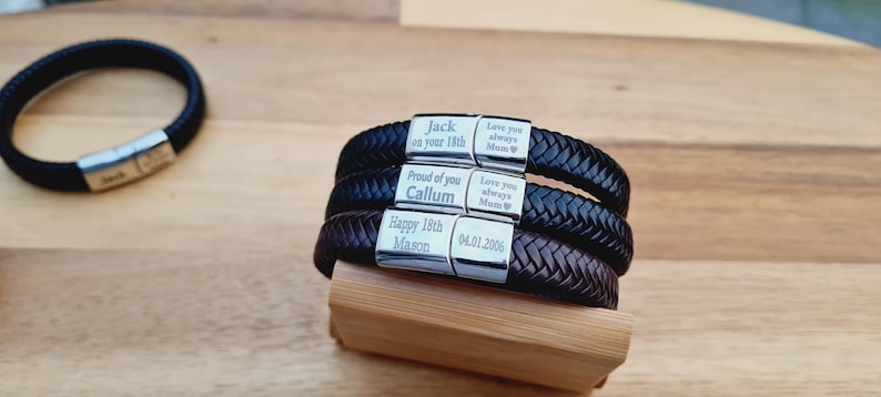 May include: Braided leather bracelets in black and brown with silver clasps. The clasps are engraved with personalized messages and dates. One bracelet is separate, while the others are stacked on a wooden block.