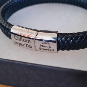 May include: A black braided leather bracelet with a silver clasp, presented in a black gift box. The clasp is engraved with the text "Callum on your 21st" and "Love Nan & Grandad". The bracelet is a stylish accessory.
