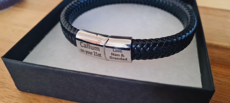 May include: A black braided leather bracelet with a silver clasp, presented in a black gift box. The clasp is engraved with the text "Callum on your 21st" and "Love Nan & Grandad". The bracelet is a stylish accessory.