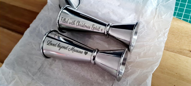 Op de afbeelding: Twee zilveren maatbekers met gegraveerde berichten. De ene beker heeft de tekst "Filled with Christmas Spirit x" en de andere "Loved beyond Measure".