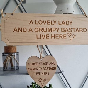 May include: A wooden sign with the text "A Lovely Lady and a Grumpy Bastard Live Here" with two small hearts. A smaller heart-shaped wooden sign hangs below with the same text.