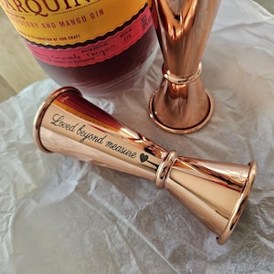 Op de afbeelding: Een koperen jigger met de inscriptie "Loved beyond measure" en een klein hartje. De jigger is een bartool die wordt gebruikt om vloeistoffen af te meten.