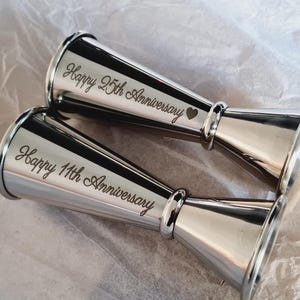 May include: Two silver measuring cups with engraved messages. The first cup reads "Happy 25th Anniversary" and the second cup reads "Happy 11th Anniversary".