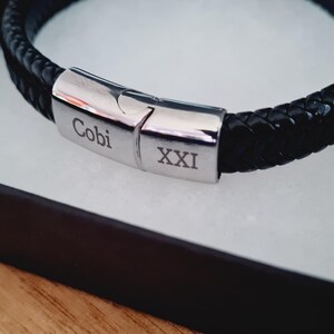 May include: A black braided leather bracelet with a silver clasp, presented in a black gift box with a white interior. The clasp is engraved with the name "Cobi" and the Roman numeral "XXI". The bracelet is displayed on a wooden surface.