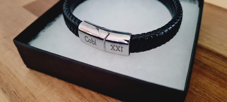 May include: A black braided leather bracelet with a silver clasp, presented in a black gift box with a white interior. The clasp is engraved with the name "Cobi" and the Roman numeral "XXI". The bracelet is displayed on a wooden surface.