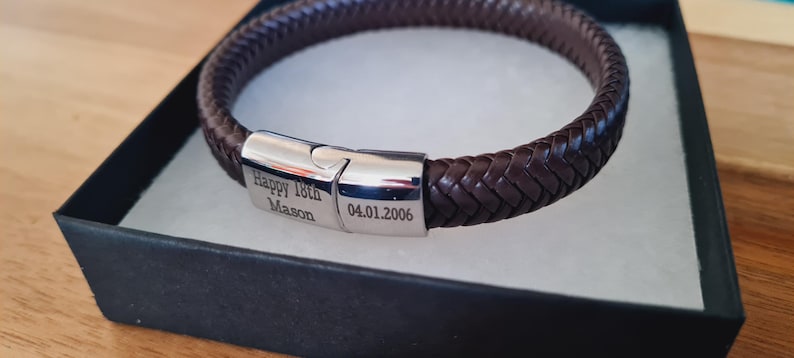 May include: A brown braided leather bracelet with a silver clasp, presented in a black gift box. The clasp is engraved with "Happy 18th Mason" and the date "04.01.2006". The bracelet is approximately 8 inches in circumference.