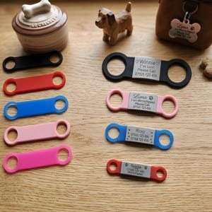 May include: A collection of colorful dog collar tags in black, red, blue, pink, and fuchsia. Some tags have engraved text with names and phone numbers. Other items include a dog treat, a dog-shaped figurine, and a dog treat jar.