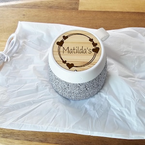 May include: A portable wireless speaker with a grey fabric base and a white plastic top. The top features a circular bamboo inlay engraved with the text 'Matilda's' surrounded by a decorative heart border. A white USB charging cable is included.
