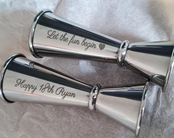 May include: Two silver jigger shot glasses with engraved text. The first jigger says "Let the fun begin" with a heart symbol. The second jigger says "Happy 18th Ryan".