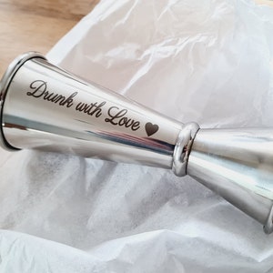 Op de afbeelding: Een zilveren cocktailjigger met de inscriptie "Drunk with Love" en een klein hartje.