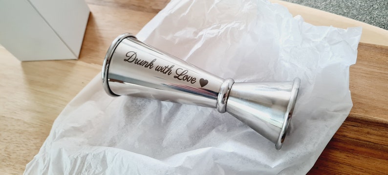Op de afbeelding: Een zilveren cocktailjigger met de inscriptie "Drunk with Love" en een klein hartje.