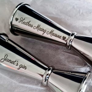Op de afbeelding: Twee zilveren cocktailmaatbekers met gegraveerde tekst. De eerste maatbeker zegt "Heather's Mixing Measure" met een hart aan beide kanten van de tekst. De tweede maatbeker zegt "Janet's gin" met een hart boven de tekst.