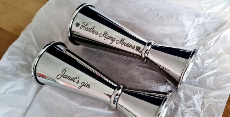 Op de afbeelding: Twee zilveren cocktailmaatbekers met gegraveerde tekst. De eerste maatbeker zegt "Heather's Mixing Measure" met een hart aan beide kanten van de tekst. De tweede maatbeker zegt "Janet's gin" met een hart boven de tekst.