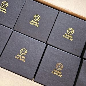 May include: A close-up shot of several black square boxes arranged in a row. Each box has the words "Bespoke Engraving" printed in gold, along with a circular logo. The boxes are inside a larger cardboard container.