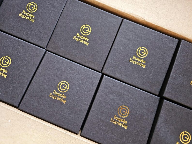 May include: A close-up shot of several black square boxes arranged in a row. Each box has the words "Bespoke Engraving" printed in gold, along with a circular logo. The boxes are inside a larger cardboard container.