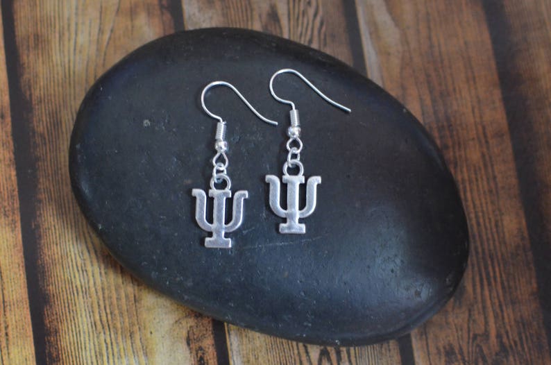 May include: A pair of silver earrings with a psychology symbol charm. The earrings are hanging from a black stone.