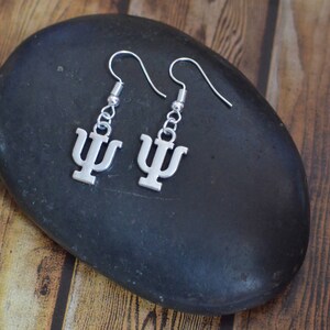 May include: A pair of silver earrings with a Greek letter psi charm. The earrings are hanging from a black stone.