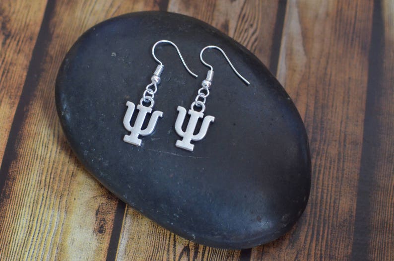 May include: A pair of silver earrings with a Greek letter psi charm. The earrings are hanging from a black stone.