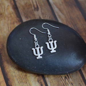 May include: A pair of silver earrings with a Greek letter psi charm. The earrings are on a black stone background.