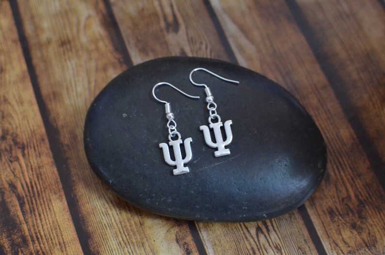 May include: A pair of silver earrings with a Greek letter psi charm. The earrings are on a black stone background.