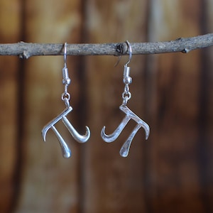 Puede incluir: Un par de pendientes plateados con la forma del símbolo matemático pi (π). Los pendientes cuelgan de ganchos plateados, sobre un fondo de madera borroso.