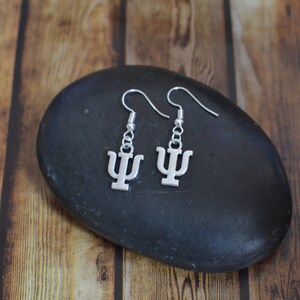 May include: Silver earrings with a Greek letter psi charm. The earrings are hanging from a black stone.