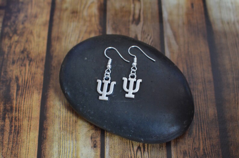 May include: Silver earrings with a Greek letter psi charm. The earrings are hanging from a black stone.