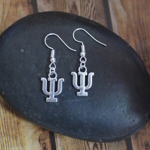 May include: A pair of silver earrings with a psychology symbol charm. The earrings are hanging from a black stone.