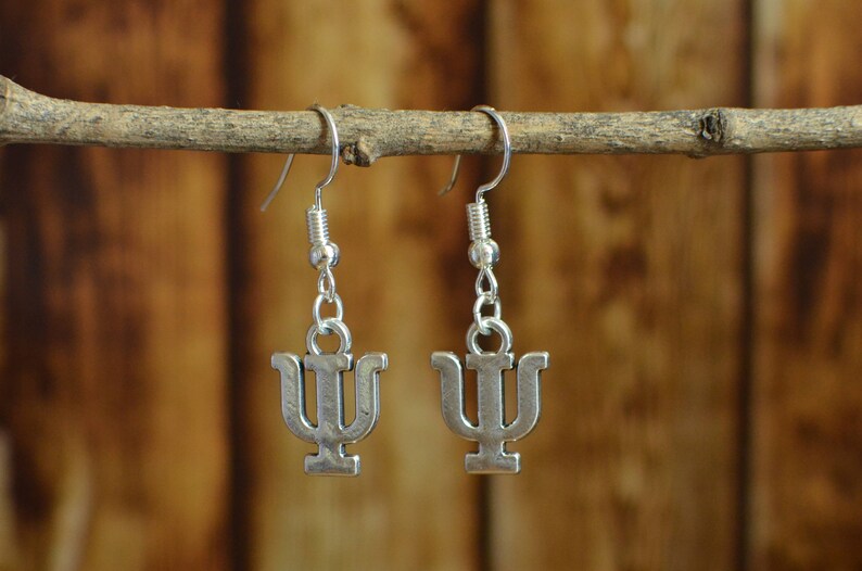 May include: A pair of silver earrings with a Greek letter psi charm. The earrings are hanging from a wooden branch.