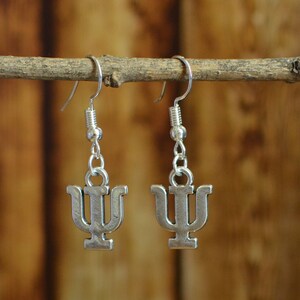 May include: A pair of silver earrings with a Greek letter psi charm. The earrings are hanging from a wooden branch.