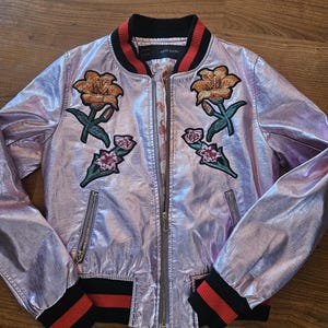 May include: A pink metallic bomber jacket with black and red striped trim. The jacket features embroidered floral designs on the front.