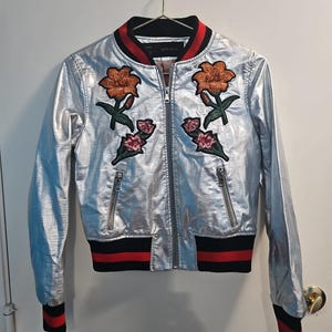 May include: Silver bomber jacket with orange and pink floral embroidery. The jacket has a black collar with red stripes, black cuffs with red stripes, and two front pockets. The jacket is hanging on a white wall.