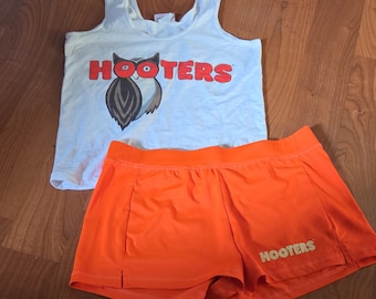 May include: A white sleeveless tank top featuring the red text HOOTERS and an owl graphic on the front, paired with bright orange athletic shorts that have the word HOOTERS printed in a light yellow color on the lower right leg area.