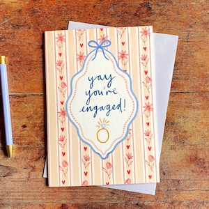 Engagement greeting card shown on wooden backdrop. Card reads yay you're engaged inside of a blue ribbon frame with florals surrounding