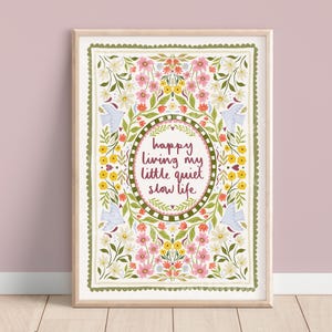 May include: A framed art print with a floral design and the text "happy living my little quiet slow life." The artwork features a white background with pink, yellow, and green floral elements, and two blue bird illustrations. The frame is a light wood colour.