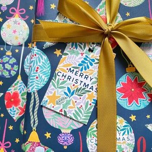 May include: A navy blue gift wrap with a gold ribbon and bow. The wrapping paper features a repeating pattern of colorful ornaments with floral designs. A gift tag with the text "Merry Christmas" is attached to the ribbon.