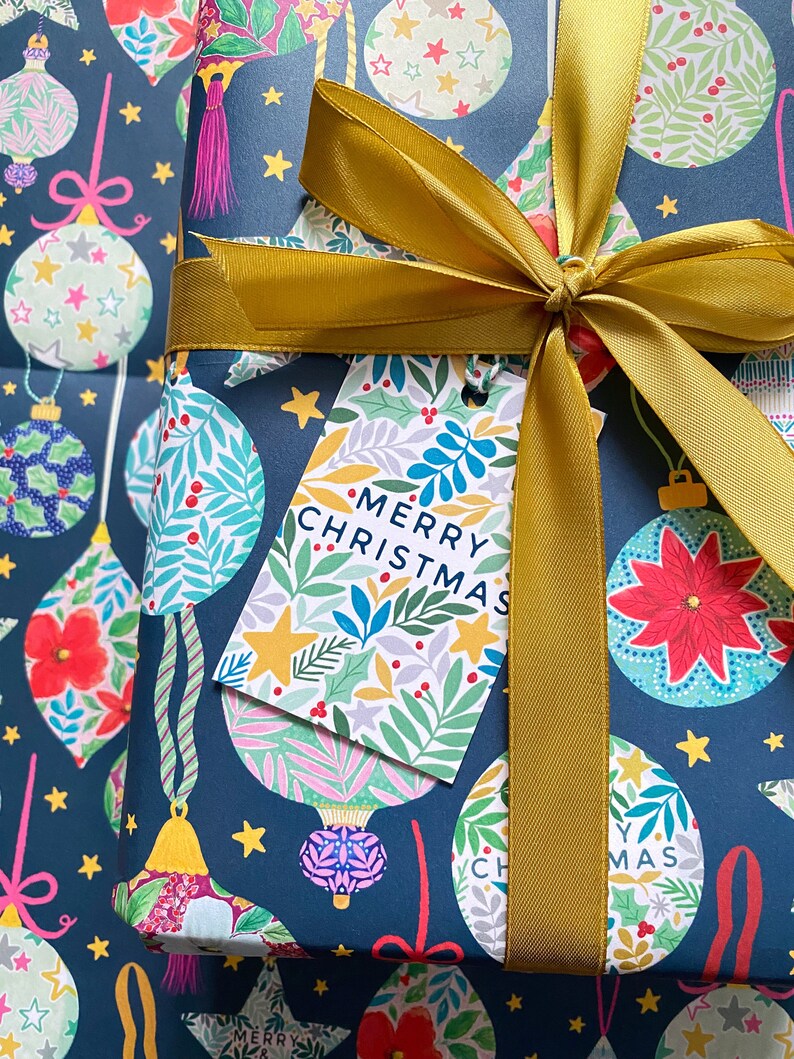May include: A navy blue gift wrap with a gold ribbon and bow. The wrapping paper features a repeating pattern of colorful ornaments with floral designs. A gift tag with the text "Merry Christmas" is attached to the ribbon.