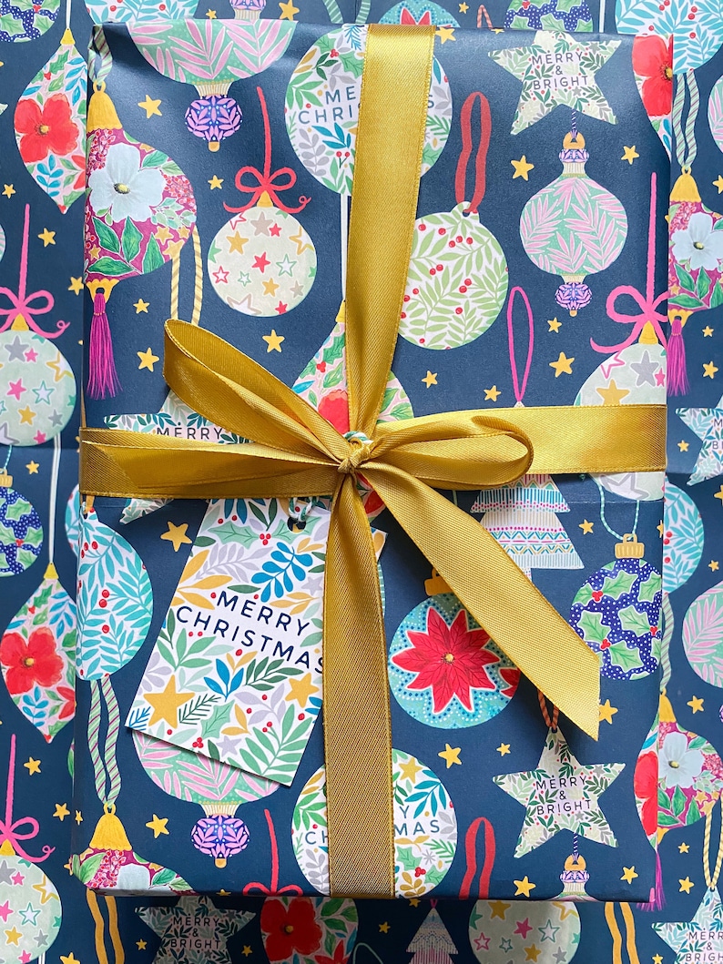 May include: A blue gift wrap with a gold ribbon and a gift tag that says "Merry Christmas". The wrap features a repeating pattern of colorful ornaments, stars, and the words "Merry and Bright".