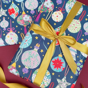 May include: Blue wrapping paper with a gold ribbon. The paper features a repeating pattern of colorful Christmas ornaments with a floral design. The text "Merry & Bright" is printed on one of the ornaments.