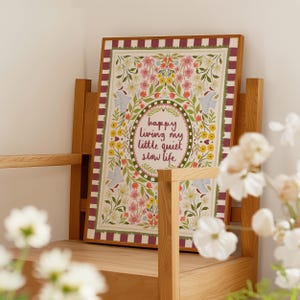 May include: A rectangular framed print featuring a maroon and white striped border. The center displays the text "happy living my little quiet slow life" surrounded by a decorative oval frame, colorful floral illustrations, and four light blue birds.