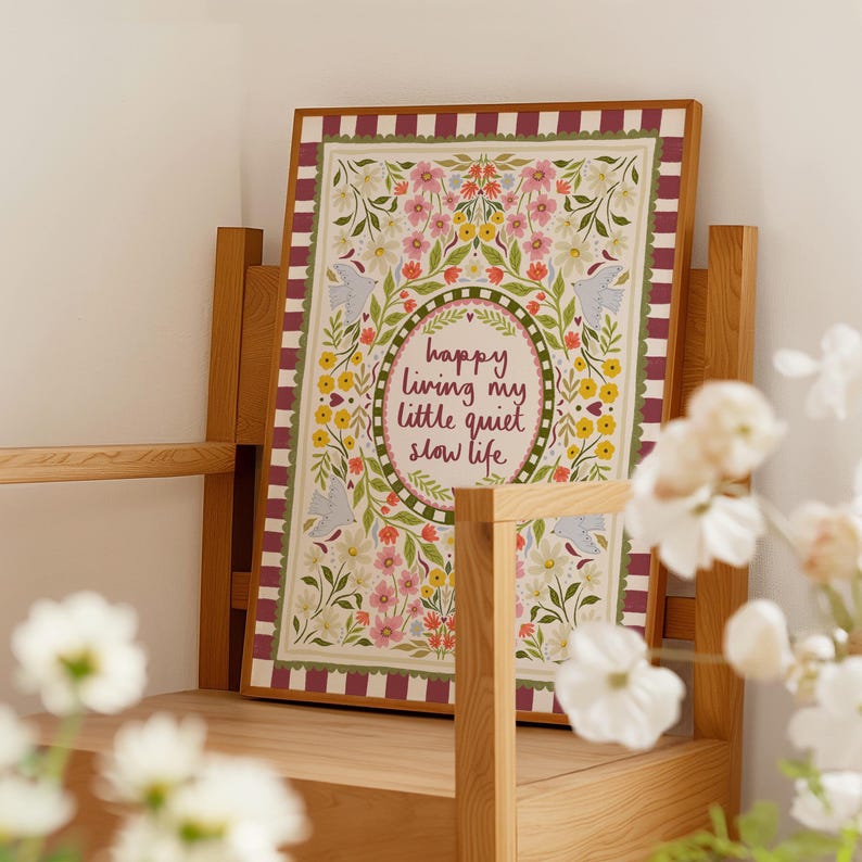 May include: A rectangular framed print featuring a maroon and white striped border. The center displays the text "happy living my little quiet slow life" surrounded by a decorative oval frame, colorful floral illustrations, and four light blue birds.