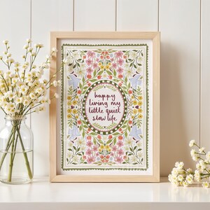 May include: A rectangular wooden frame containing a print with a floral border in shades of pink, yellow, and green. The center features an oval frame enclosing the text "happy living my little quiet slow life" in dark red cursive lettering.