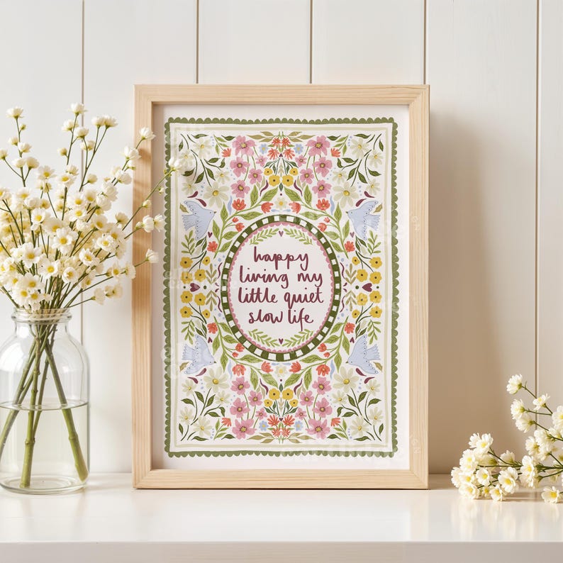 May include: A rectangular wooden frame containing a print with a floral border in shades of pink, yellow, and green. The center features an oval frame enclosing the text "happy living my little quiet slow life" in dark red cursive lettering.
